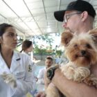 Mulher com luvas e jaleco conversa com homem que segura um cachorro no colo