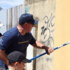 Imagem mostra homem usando boné e manuseando rolo com tinta amarela em uma parede. Ao seu lado, estão uma mulher de boné segurando um rolo e um menino, também de boné