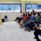 Mulher fala em microfone em púlpito, intérprete de libras ao lado dele, ambos estão em frente a plateia