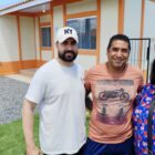 Dois homens e uma mulher posam para foto em pé, na frente de uma casa em tons de laranja, com gramado na frente. A mulher sorri e exibe a chave de uma casa