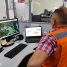 Homem com colete laranja da Defesa Civil está sentado observando dois monitores, de costa para a câmera