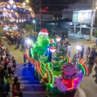 Imagem aérea mostra carro alegórico enfeitado com grande jacaré verde usando gorro de natal em desfile acompanhando por várias pessoas