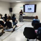 Mulher está em pé à frente de sala e perto de telão enquanto pessoas sentadas acompanham a apresentação