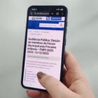 Mão de mulher usando esmalte segura celular que mostra página da Prefeitura de Joinville com título sobre audiência pública