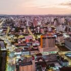 Imagem aérea de Joinville ao entardecer, com prédios, ruas iluminadas e montanhas ao fundo