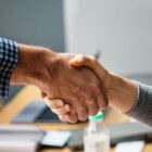 Imagem mostra o aperto entre duas mãos, uma feminina e outra masculina, com ambiente de escritório ao fundo