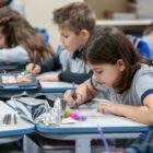 Em primeiro plano, menina está escrevendo em papel sobre a mesa. Ao fundo, outras crianças uniformizadas também estão escrevendo em suas carteiras