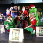 Em um palco, um homem está vestido de Papai Noel junto de sua bicicleta decorada. Ao lado dele, estão dois duendes, o jacaré Fritz, o boneco de neve, o biscoito, um papai noel vestido de azul e um homem apontando para o ciclista