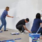Dois homens e duas mulheres estão com rolos de tinta nas mãos pintando uma parece branca com pichação vermelha. Há baldes de tinta, bandejas e materiais de pintura no entorno
