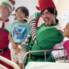 Dois homens vestidos de duendes sorriem para a foto. Um deles está com as mãos no rosto de uma criança, que sorri também. Outra criança ao lado olha para ele também sorrindo