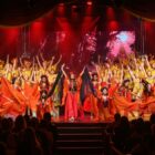 Dezenas de pessoas estão em um palco, em pé, com os braços levantados vibrando. Eles vestem figurinos laranja e vermelhos