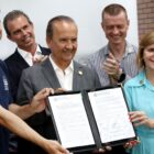 Dois homens e uma mulher sorriem para foto enquanto seguram um documento. Ao fundo, outras pessoas também sorriem e aplaudem