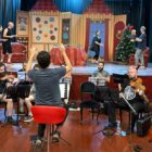 Músicos de orquestra ensaiam em frente ao palco, no palco há atores e cenário