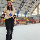 Em pista de patinação no gelo, duas mulheres patinam perto de uma criança, todas com equipamentos de segurança. Ao fundo, outras pessoas também patinam no gelo