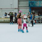 Em primeiro plano, menina e mulher usam patins e estão de mãos dadas na pista ampla. Ao fundo, outras pessoas aparecem patinando e observando