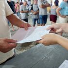 Imagem mostra um documento sendo repassado de uma pessoa para a outra. Apenas os braços aparecem