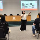 Mulher com cabelo castanho calça preta e blusa branca fala ao microfone em pé para plateia sentada