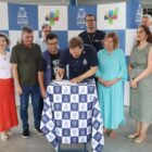 Homens e mulheres estão em pé, em frente a um painel com a marca da Prefeitura e da Secretaria da Saúde. Eles observam um homem de camisa azul assinar um documento