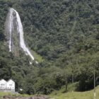 Em área repleta de montanhas e vegetação, uma cachoeira com grande queda d'aágua e um prédio branco se destacam