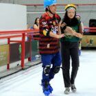 Rapaz, deficiente visual, com capacete e joelheira, patina em pista de patinação no gelo, acompanhada por moça