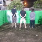 Dois homens e mulher estão de costas para a câmera, todos com os braços para trás, ao lado de um dois guardas municipais fardados