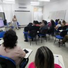 Mulher loira está em pé, ao lado de telão, fazendo apresentação para outras pessoas sentadas em uma sala