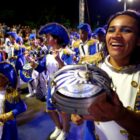 Bateria de escola de samba reúne pessoas com figurino azul, branco e dourado. Em primeiro plano, uma mulher sorri enquanto toca um pandeiro