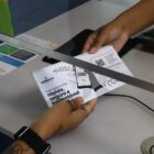 Imagem mostra duas mãos de pessoas diferente segurando um mesmo carnê por baixo do vidro de um guichê de atendimento