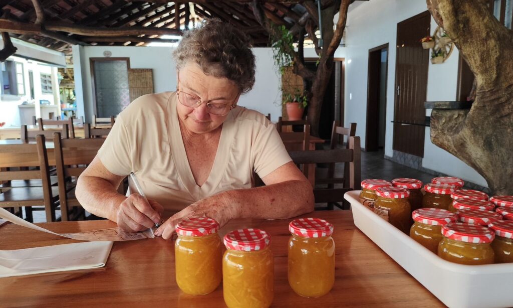 Mulher idosa escreve em uma etiqueta. À sua frente, estão potes de geleia amarela