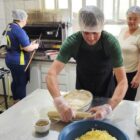 Um homem e duas mulheres estão em uma cozinha e usam toucas de proteção na cabeça. Ele está embalando uma forma de salgado com um plástico filme, enquanto a mulher mais velha observa e a mais nova acompanha uma receita no forno, de costas para a câmera