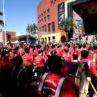 Dezenas de pessoas estão uniformizadas com roupas vermelhas e tocam instrumentos em frente ao Centreventos Cau Hansen