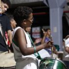 Menino negro toca instrumento de percussão. Ao fundo, outras pessoas tocam instrumentos na bateria