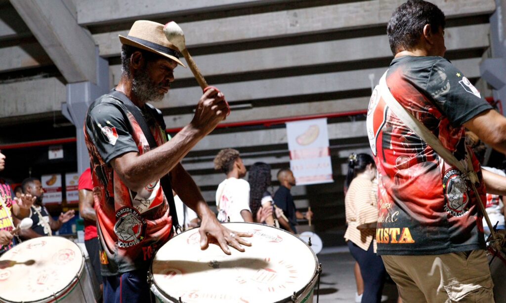 Homem negro usando chapéu veste camisa preta, branca e vermelha enquanto toca instrumento de percussaõ