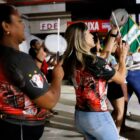 Uma mulher negra e outra loira vestem camisa preto, branco e vermelha estão em pé, tocando instrumento