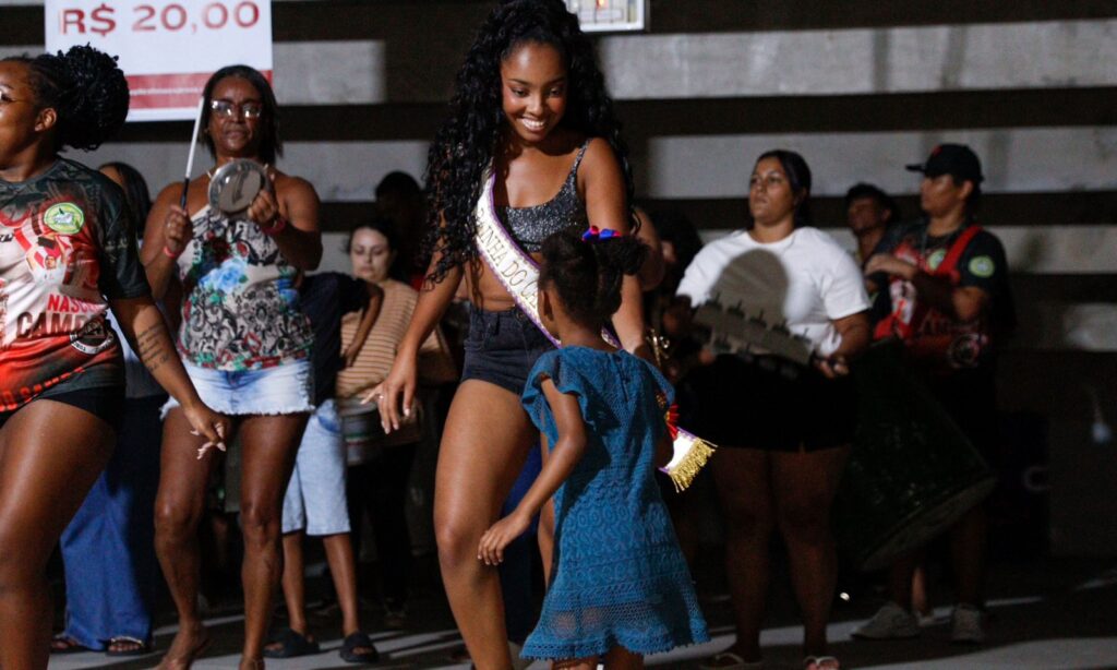 Mulher negra com faixa de rainha do Carnaval sorri para menina de vestido enquanto as duas dançam