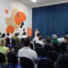 Pessoas sentadas em cadeira em auditório ouvem mulher em pé com microfone na mão