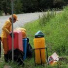 Mulher com uniforme e boné amarelos verifica recipiente com lixo às margens de uma estrada de terra