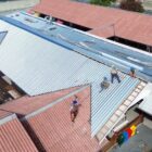 Imagem mostra trabalhadores utilizando equipamentos de proteção individual trabalhando em cima do telhado de uma unidade escolar