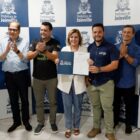 Uma mulher e seis homens estão em pé. Ela está no centro e segura um documento enquanto sorri. Os demais aplaudem