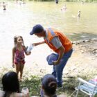 Homem com colete laranja e boné orienta criança na beira de rio, ele tem material informativo nas mãos
