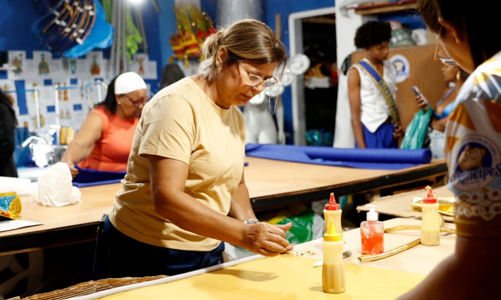 Mulher loira de óculos está próxima a uma mesa, onde manuseia cola e outros materiais para produzir fantasia