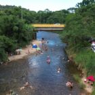 Imagem aérea mostra um rio com banhistas, vegetação no entorno e ponte ao fundo da imagem