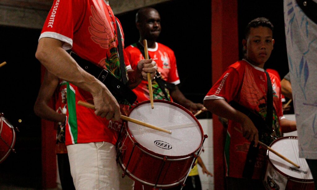 Dois homens e um menino estão com camisas vermelhas e tocam instrumentos de percussão