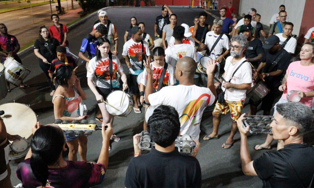 Homens e mulheres estão em pé, em espaço externo, e tocam diferentes instrumentos