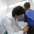 Mulher de cabelo curto usando jaleco branco aplica vacina no quadril de um menino negro que está em pé e de costas na sala de vacinação