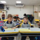 Sala de aula está repleta de crianças uniformizadas. Dois meninos estão em foco nas carteiras da frente. Um deles sorri para a câmera, enquanto o outro olha para a atividade do colega.