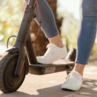 Imagem mostra parte de um patinete e as pernas de uma pessoa, uma no chão e outra sobre o veículo