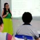 Mulher de óculos com saia e blusa verdes está à frente de sala de aula, perto de quadro, enquanto dois alunos sentados de costas para a câmera a observam