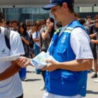 Agente de trânsito com boné e colete distribui panfleto para alunos em escola