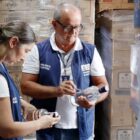 Homem e mulher estão vestidos com coletes azuis, da Vigilância Sanitária, e cada um observa uma garrafa de bebida transparente